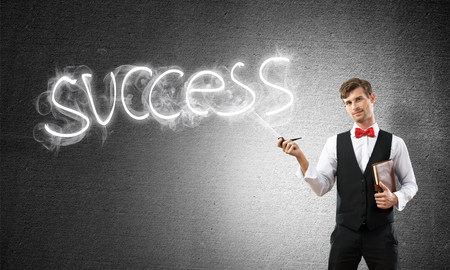 Horizontal shot of confident and young businessman in smart-casual wear smoking pipe while standing against gray dark wall and success word made from smoke on background.の写真素材