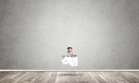 Young little boy keeping eyes closed and looking concentrated while meditating on cloud in the air with gray concrete wall on background.の写真素材