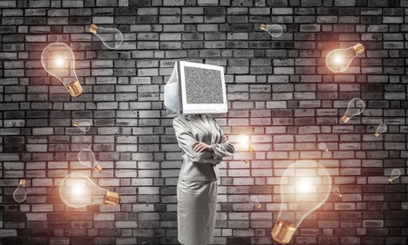 Business woman in suit with monitor instead of head keeping arms crossed while standing among flying lightbulbs and with gray dark wall on background.の写真素材