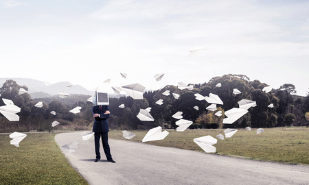 Businessman in suit with monitor instead of head keeping arms crossed while standing on the road among flying paper planes with beautiful landscape on background.の写真素材