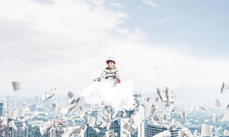 Young little boy keeping eyes closed and looking concentrated while meditating on cloud among flying papers with cityscape view on background.の写真素材
