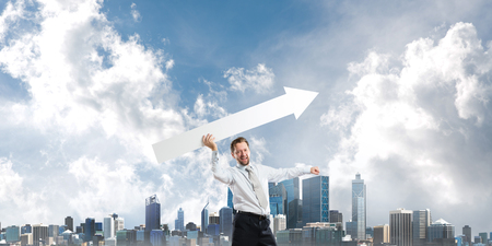 Conceptual image of attractive businessman launching huge white arrow to the sky while standing against modern cityscape view on background.の写真素材