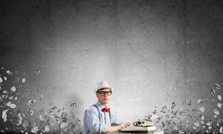 Young man writer in hat and eyeglasses using typing machine while sitting at the table among flying letters and against gray concrete wall on background.の写真素材