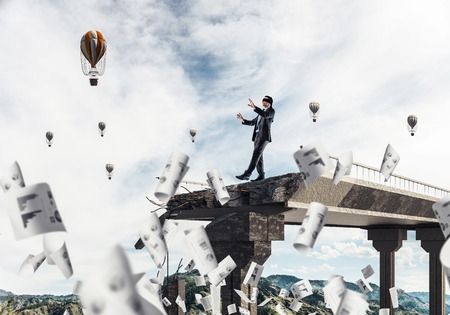 Businessman walking blindfolded among flying documents on concrete bridge with huge gap as symbol of hidden threats and risks. Flying balloons and nature view on background. 3D rendering.の写真素材