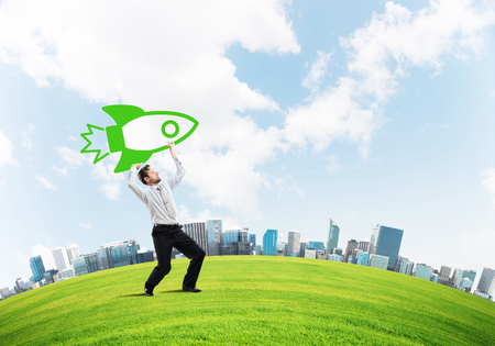 Horizontal shot of successful and young businessman standing on green meadow and throwing huge rocket in the air with cloudy skyscape view on background.の写真素材