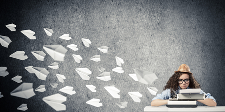 Young and beautiful woman writer in hat and eyeglasses using typing machine while sitting at the table among flying paper planes and against gray concrete wall on background.の写真素材