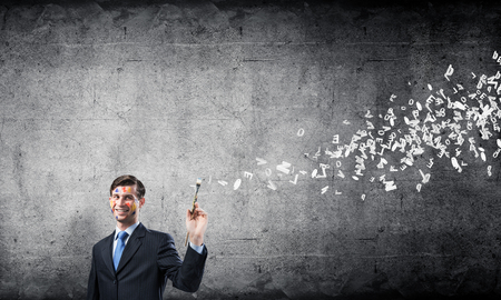Conceptual image of young and successful businessman in black suit holding paintbrush in hand while standing with flying letters and dark gray wall on background.の写真素材