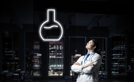 Horizontal shot of young confident doctor in white medical uniform interracting with glowing vial symbol whie standing against night cityscape view on background.の写真素材