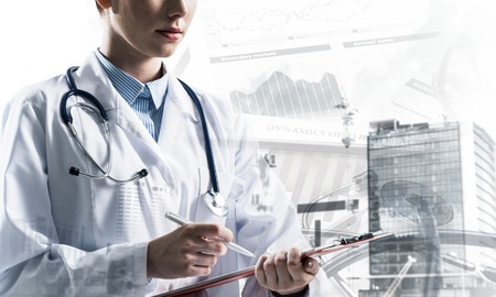 Portrait of young female doctor in white sterile suit holding notebook in hands while standing against modern city view and medical equipment on background. Double exposureの写真素材