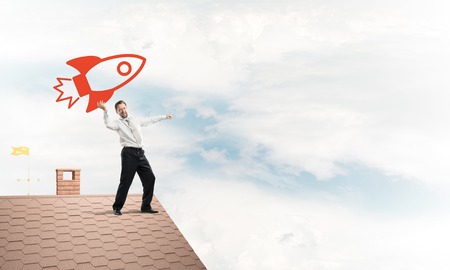 Successful and young businessman standing on the top of brick roof and throwing huge red rocket in the air with cloudy skyscape view on background.の写真素材