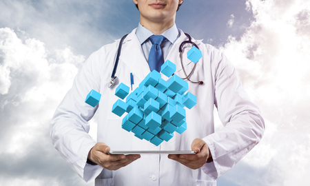 Confident and professional doctor in medical uniform presenting set of multiple cubes above tablet in his hands while standing with blue skyscape on background.の写真素材