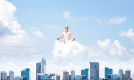 Woman in white clothing keeping eyes closed and looking concentrated while meditating on cloud in the air with cityscape view on background.の写真素材