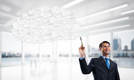 Conceptual image of young and successful businessman in black suit holding paintbrush in hand and smiling while standing with chatting cloud inside modern bright office.の写真素材