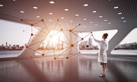 Horizontal shot of young female doctor in white medical uniform working with social network structure while standing inside white hospital office. Medical industryの写真素材