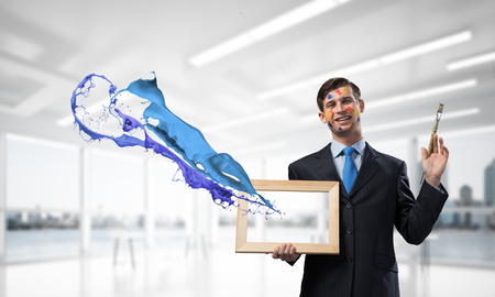 Horizontal shot of cheerful and young businessman in black suit gesturing and smiling while standing near blue liquid splash inside bright modern office.の写真素材