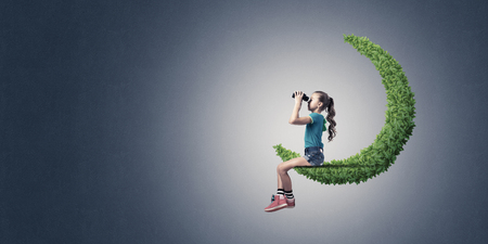 Cute kid girl sitting on green moon and looking in binocularsの写真素材