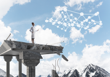 Young woman doctor in white uniform standing at end of broken bridge and examining social media network structure. Medical industry concept. Landscape view on backgroundの写真素材