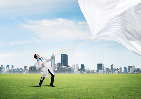 Horizontal shot of young doctor in white medical uniform pulling white fabric while standing on green meadow with cityscape view on backgroundの写真素材