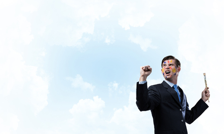 Horizontal shot of cheerful and young businessman in black suit gesturing and smiling while standing against blue cloudy skyscape on background.の写真素材