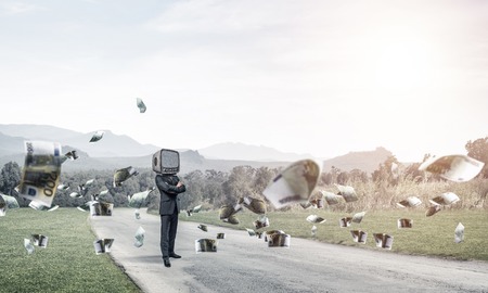 Businessman in suit with TV instead of head keeping arms crossed while standing on the road among flying euro banknotes with beautiful landscape on background.の写真素材