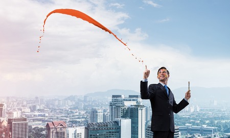 Conceptual image of ambitious and creative businessman in black suit holding paintbrush in hand and looking away while standing with orange splash against cityscape view on background.の写真素材