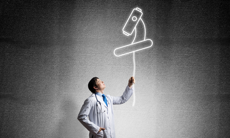 Young male doctor in white uniform interracting with glowing microscope icon in the air while standing against gray dark wall on background.の写真素材