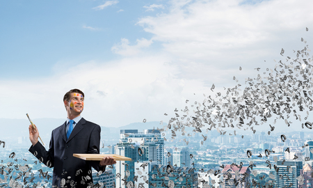 Successful and confident businessman keeping paintbrush in his hand and looking away while standing against cityscape view with flying letters on backgroundの写真素材
