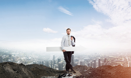 Horizontal shot of young businessman holding white arrow which is pointing sideways while standing against cityscape view and cloudy sky on background.の写真素材