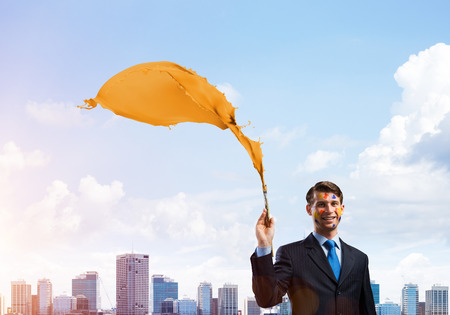 Young and successful businessman in black suit holding paintbrush in hand and smiling while standing with orange liquid splash with against modern cityscape view on background.の写真素材
