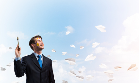 Conceptual image of young businessman in suit holding paintbrush in his hand and smiling while standing against blue cloudy skyscape with flying paper planes on background.の写真素材