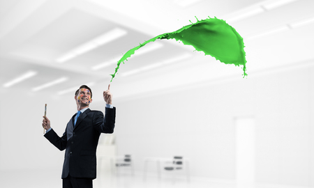 Young and successful businessman in black suit holding paintbrush in hand and smiling while standing with colourful liquid splash inside modern bright office.の写真素材