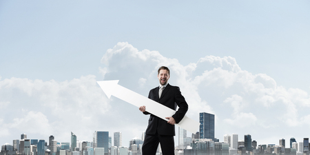 Conceptual image of young businessman in black suit showing aside with big white arrow in his hands...