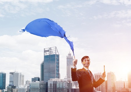 Successful businessman in black suit gesturing and smiling while standing with blue coloured splash against cityscape view on background.の写真素材