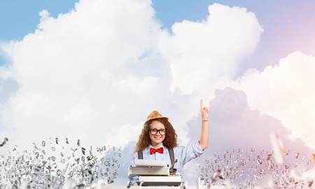 Young and beautiful woman writer in hat and eyeglasses using typing machine and pointing upside...