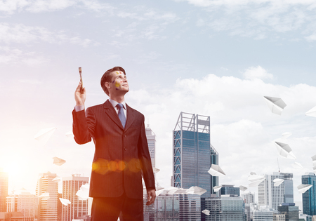 Studio shot of successful and confident keeping paintbrush in his hand and looking away while standing against cityscape view with flying paper planes on backgroundの写真素材