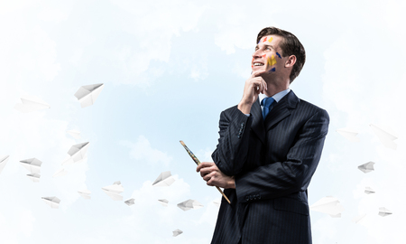 Portrait of young thoughtful man holding paintbrush in his hand and looking away while standing against blue cloudy skyscape with flying paper planes on background.の写真素材