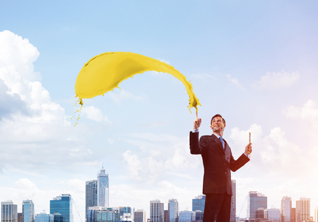 Horizontal shot of young and happy businessman in black suit holding paintbrush in his hand while standing with yellow liquid splash against cityscape view on background.の写真素材