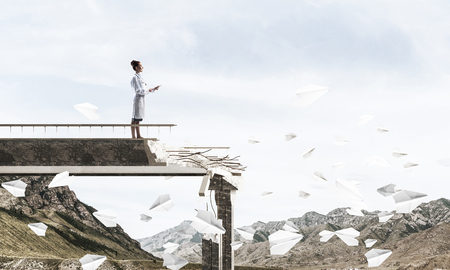 Professional medical industry employee using tablet while standing among flying paper planes at the edge of broken bridge with landscape view on background. Medical industry conceptの写真素材