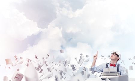 Young man writer in hat and eyeglasses using typing machine and pointing upside while sitting at the table among flying papers with cloudy skyscape on background.の写真素材