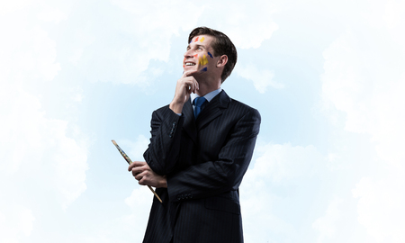 Portrait of young thoughtful man holding paintbrush in his hand and looking away while standing against blue cloudy skyscape on background.の写真素材