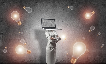 Business woman in suit with laptop instead of head keeping arms crossed while standing among flying lightbulbs and with gray dark wall on background.の写真素材