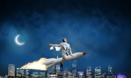 Conceptual image of young businessman in suit flying on rocket with night cityscape and blue sky on background.の写真素材