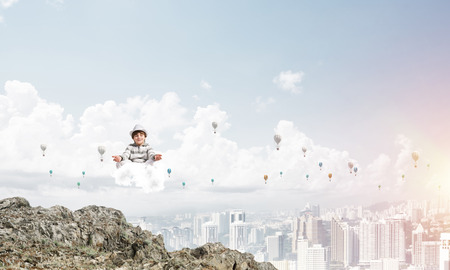 Young little boy keeping eyes closed and looking concentrated while meditating on cloud in the air with city view on background.の写真素材