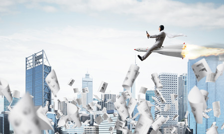 Conceptual image of young and happy businessman in suit flying on rocket among flying papers with modern cityscape with skyscrapers and blue sky on background.の写真素材
