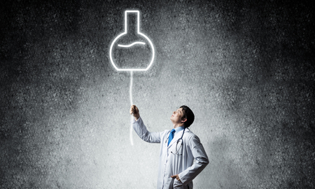 Young confident doctor in white medical uniform interracting with glowing vial symbol whie standing against dark gray wall on background.の写真素材