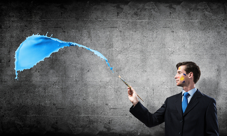 Successful businessman in black suit gesturing and smiling while standing with blue coloured splash against gray concrete wall on background.の写真素材