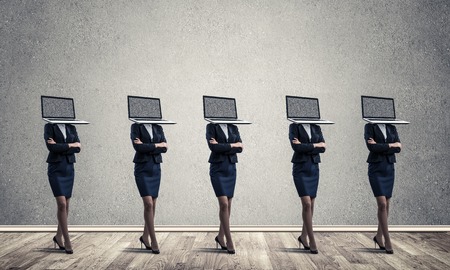 Business women in suits with laptops instead of their heads keeping arms crossed while standing in a row in empty room with gray wall on background.の写真素材
