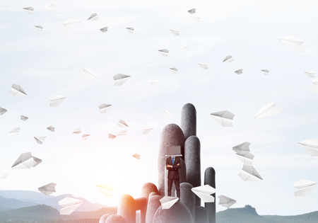 Businessman in suit with laptop instead of head keeping arms crossed while standing on the top of stone column among flying paper planes with beautiful skyscape on background. 3D rendering.の写真素材