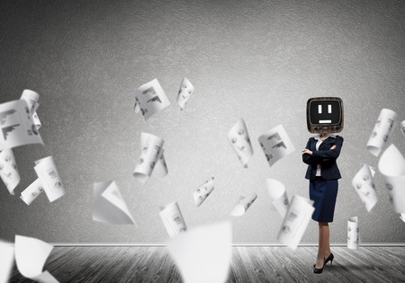 Business woman in suit with old TV instead of head keeping arms crossed while standing among flying papers inside empty room with gray dark wall on background.の写真素材