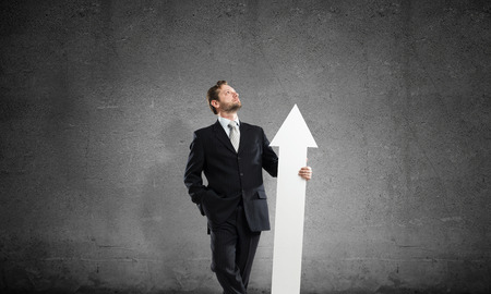 Confident and successful businessman in suit pointing upside with huge white arrow in his hands while standing against dark gray wall on backgroundの写真素材